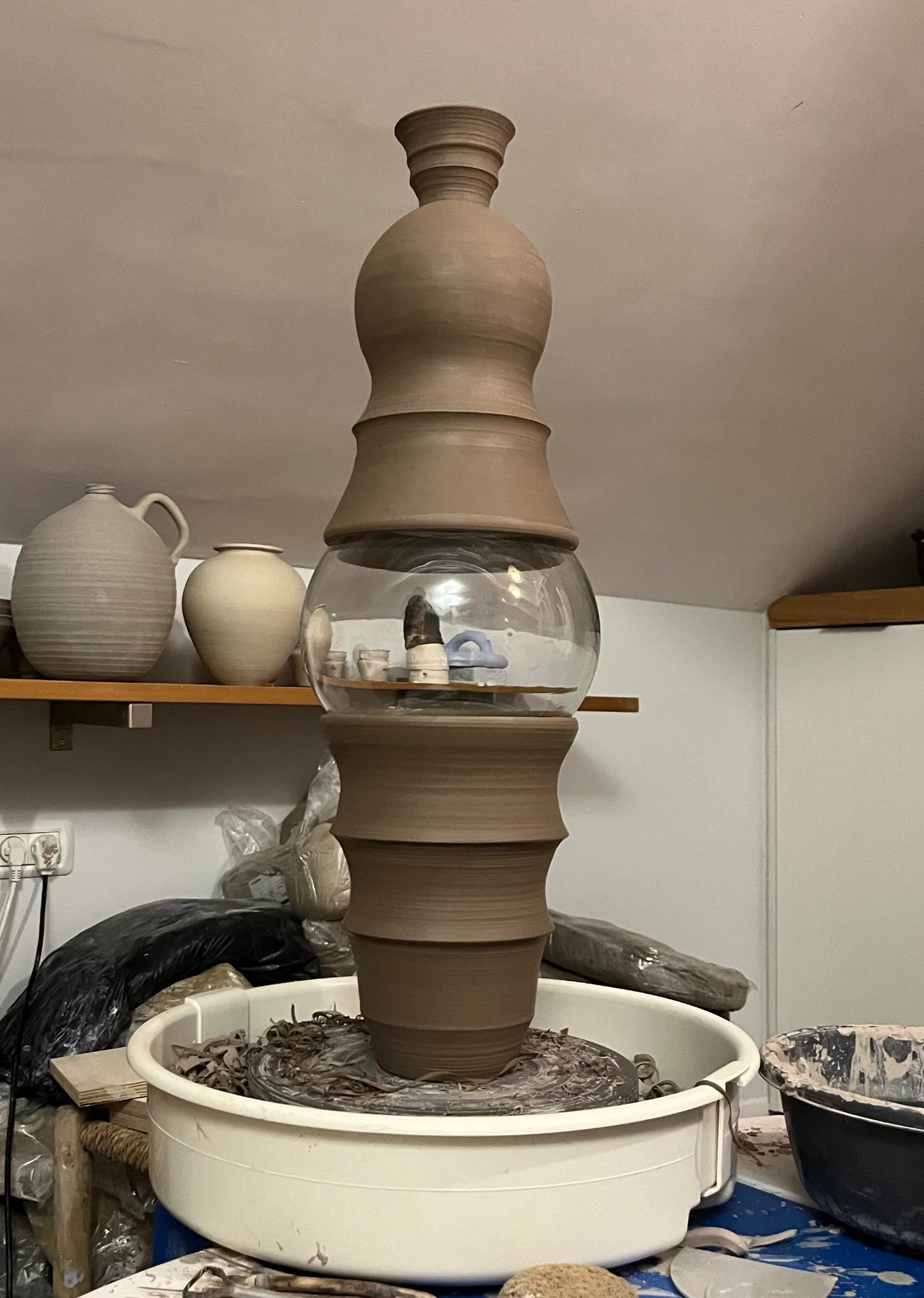 A tall stacked clay and glass form on the wheel in the studio